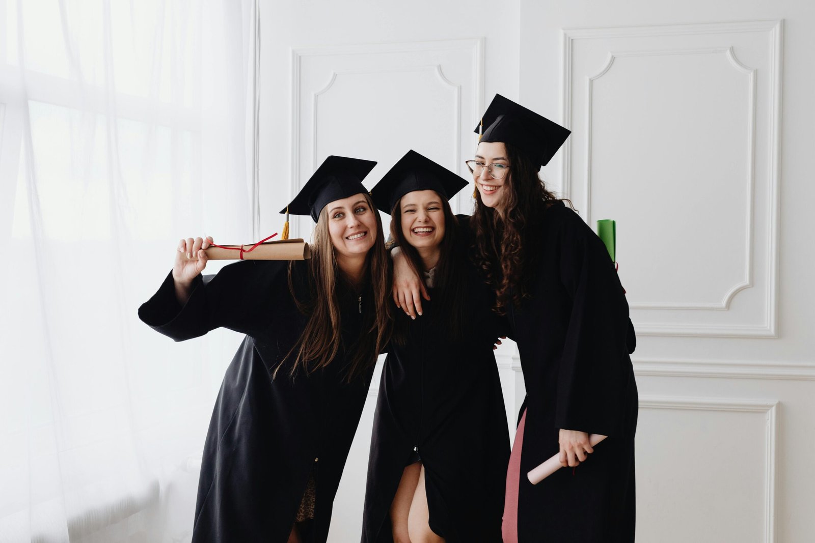 Three smiling graduates in caps and gowns celebrating indoors together.